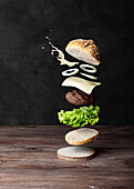 Ingredients of delicious burger falling from above on wooden surface against black background