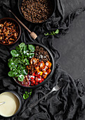 Top view of stewpan with lentils near salad with pumpkin and bell pepper decorated with green basil leaves on black background near salad dressing