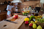 Soft focus of assorted fruits and ripe tomato placed near cutting board and knife on table near woman taking utensil from rack during salad preparation in kitchen at home