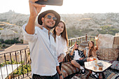 Group of young diverse friends taking selfie on cellphone while drinking cocktails on terrace bar in Cappadocia, Turkey
