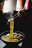 Hand of unrecognizable woman picking noodles with chopsticks from bowl of savory Asian soup in cafe