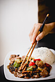 Hand of anonymous female using chopsticks to pick piece of beef in delicious oyster sauce from plate in restaurant