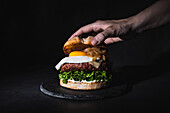 Crop unrecognizable person putting bun on burger with egg and patty served on slate board on black background in studio