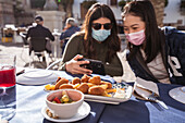 Positive Asian woman with female friend in medical masks watching video on cellphone while sitting at table in outdoor cafeteria