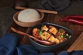 Nahaufnahme von Mala Tofu, einem chinesischen veganen Gericht, begleitet von einer Schüssel Reis und einer japanischen Teekanne auf einem mit Stoffen dekorierten Holztisch