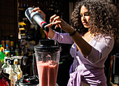 Zufriedene junge ethnische Barkeeperin in Freizeitkleidung gibt süßen Sirup in einen Mixer mit Beeren-Smoothie, während sie in einer sonnigen Bar im Freien Getränke mixt