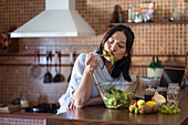 Hungry young Asian female with long dark hair in casual clothes eating appetizing healthy salad with closed eyes while leaning on counter in kitchen
