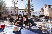 Positive Asian woman with female friend in medical masks watching video on cellphone while sitting at table in outdoor cafeteria