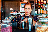Content female bartender pouring alcohol drink in metal shaker while preparing refreshing cocktail at counter in bar