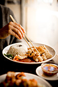 Hand einer Frau, die am Tisch sitzt und mit Stäbchen chinesisches gebratenes Fischgericht von einem weißen Keramikteller isst