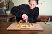 Focused Asian female cutting tasty baked pizza with sharp cutter served on wooden cutting board at table in light kitchen