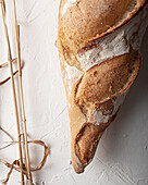 Draufsicht auf ein köstliches, frisch gebackenes, rustikales, handwerklich hergestelltes Baguette auf einer weißen Fläche mit getrockneten Weizenspitzen