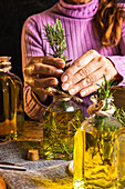 Crop faceless female in purple sweater putting herbs twigs with green leaves in essential oil glass bottle near scissors and rope with small chest on cloth at wooden table