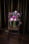 Bouquet of fresh colorful peonies and chrysanthemums in glass vase placed on weathered wooden chair near curtains in light room