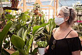 Reife Einkäuferin in Textilmaske mit Basilikum im Topf, die beim Pflücken tropischer Pflanzen im Gartengeschäft nach oben schaut