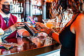 Barkeeperin mit POS-Terminal steht an der Theke mit einer anonymen Frau, die mit einer Plastikkarte für eine Bestellung in einer Bar bezahlt