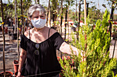 Side view of mature female shopper in textile mask picking green trees in pots in garden shop on sunny day