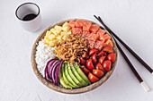 From above bowl of tasty poke dish with raw ingredients placed near chopsticks and cup of soy sauce on white plaster surface