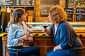 Seitenansicht von lachenden Freundinnen, die sich unterhalten und ein Glas Rotwein trinken, während sie an der Theke eines Cafés sitzen