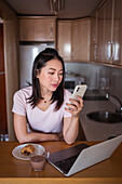 Positive Asian female text messaging on cellphone while sitting at table with netbook and food with hot drink in kitchen