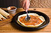 Gesichtslose Frau verteilt süße Dulce de leche auf frischem Crêpe auf schwarzem Teller auf Holztisch in Küche