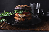Appetizing burger with sesame seeds placed on black plate near salad leaves against glass of water on dark background
