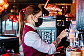Seitenansicht einer Barkeeperin mit Schutzmaske, die eine Kasse bedient und den Bildschirm eines Displays berührt, während sie in einer Bar arbeitet