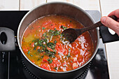 From above of crop anonymous male steering marinara sauce in pan with wooden spoon while cooking on stove in kitchen