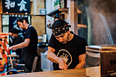 Junger Mann in Schürze kocht asiatische Gerichte, während er am Tresen einer Ramen-Bar steht
