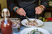 Unkenntlich gemachter männlicher Koch in schwarzer Uniform mit Armbändern serviert köstlichen Salat auf einem Teller auf dem Küchentisch