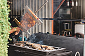 Crop faceless person standing with delicious pork ribs in hand over hot grill with firewood on terrace of light cafe