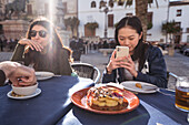 Focused Asian female taking picture of tasty shrimp dish on smartphone while sitting at table with friends during lunch in outdoor cafeteria