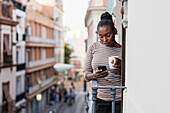 Afroamerikanische Frau mit einer Tasse Heißgetränk surft mit ihrem Handy auf dem Balkon im Internet