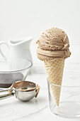 Yummy chocolate ice cream in crunchy waffle cone placed in glass bowl near pitcher and steel scoop on kitchen table