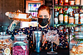 Barkeeperin mit Schutzmaske gießt alkoholisches Getränk in einen Shaker, während sie einen Cocktail zubereitet und in einer Bar während des Coronavirus arbeitet