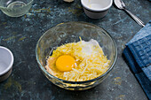 Von oben auf Glasschale mit rohem Ei und geriebener Butter für süße Waffeln mit Käse auf Tisch in heller Küche