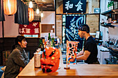 Seitenansicht einer asiatischen Frau in Freizeitkleidung, die am Tresen sitzt und sich mit einem männlichen Angestellten einer modernen Ramen-Bar unterhält