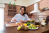 Happy young Asian female cutting fresh banana on chopping board near bowl with salad while cooking healthy food in sunlit kitchen
