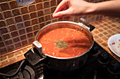 From above of unrecognizable crop female pouring aromatic spices in saucepan with hot marinara sauce made from tomatoes on stove in domestic kitchen