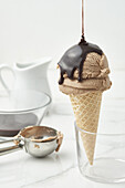 Süße Schokoladen-Ganache, die auf eine appetitliche Gelato-Kugel in einer knusprigen Waffeltüte in einer Glasschale auf einem Marmortisch in einer hellen Küche gegossen wird