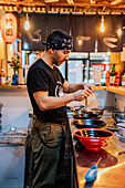 Seitenansicht eines männlichen Kochs in schwarzer Uniform und Kopftuch beim Kochen eines asiatischen Gerichts namens Ramen in einem modernen Café