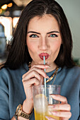 Cheerful young female in blue sweater sipping cold fizzy soft drink through straw while spending free time in cafeteria and looking at camera happily