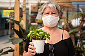 Reife weibliche Einkäuferin in Textilmaske mit Basilikum im Topf schaut in die Kamera, während sie tropische Pflanzen in einem Gartengeschäft auswählt