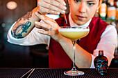 Professional young female bartender adding alcohol from skull shaped bottle with dropper into glass while preparing sour cocktail in bar