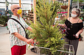 Ältere Einkäufer mit Gesichtsmasken interagieren, während sie mit einem Nadelbaum im Einkaufswagen stehen und Pflanzen in einem Gartengeschäft pflücken