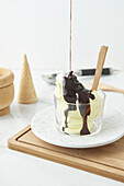 Hoher Blickwinkel auf köstlichen Schokoladensirup, der auf leckeres hausgemachtes Eis in einer Glasschüssel auf einem hölzernen Schneidebrett neben einer Waffeltüte in der Küche gegossen wird