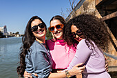 Cheerful multiethnic female friends in casual outfits embracing each other on city waterfront and looking at camera with smiles on sunny summer day