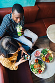 From above of anonymous female vlogger with black partner taking photo of healthy meal on cellphone in restaurant