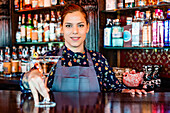 Cheerful female barkeeper with glass of refreshing vermouth standing at counter in bar and looking at camera