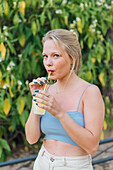 Charming female drinking cold refreshing lemonade with straw while chilling in summer garden and looking at camera